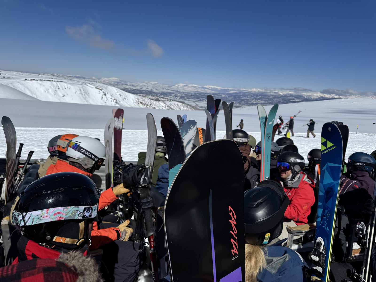 A ski crew gathered at the summit, skis and boards up, mountains behind